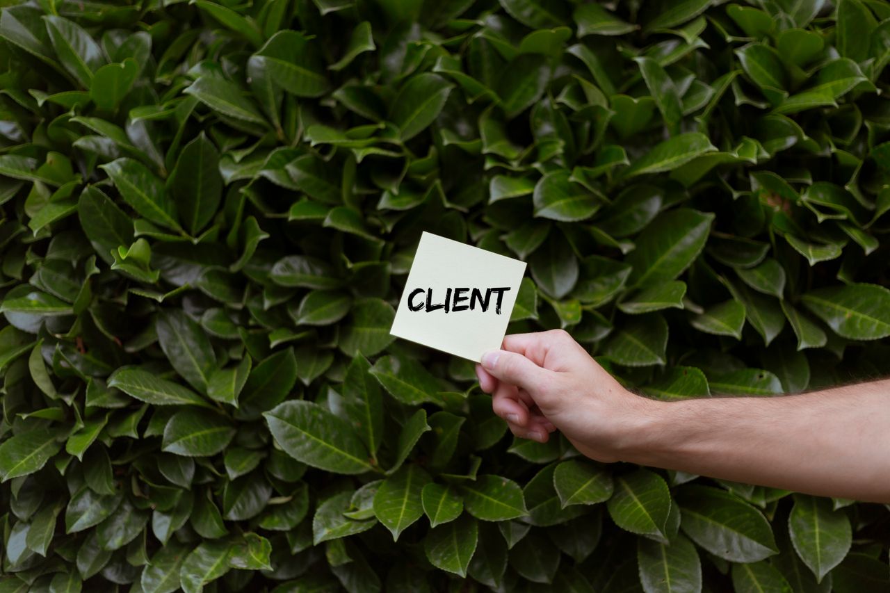 about-img-01 Close-up of a hand holding a note with the word CLIENT in front of a lush green leafy background.